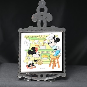 May include: Vintage cast iron trivet featuring a colorful illustration of Mickey and Minnie Mouse cooking in a kitchen. The square tile is set within a decorative, ornate frame. The trivet is designed to protect surfaces from hot cookware.