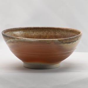 May include: A handmade ceramic bowl with a wide, rounded shape. The bowl features a gradient of colors, transitioning from a light beige rim to a warm orange-brown center, and a white base. The bowl is approximately 6 inches in diameter.