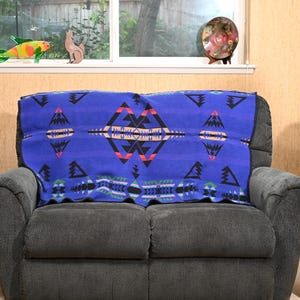 May include: A rectangular blue blanket featuring a repeating geometric pattern in black, red, orange, and white. The design includes symmetrical diamond and triangular shapes arranged horizontally across the fabric, which is draped over a gray sofa.