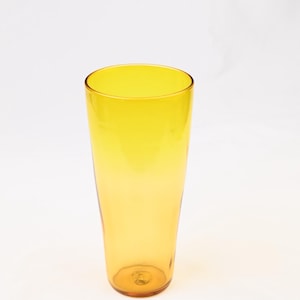 May include: A tall, tapered drinking glass made of translucent yellow glass. The vessel features a smooth, cylindrical body that gradually narrows toward the base, showcasing a vibrant, uniform golden-yellow hue throughout the entire structure.