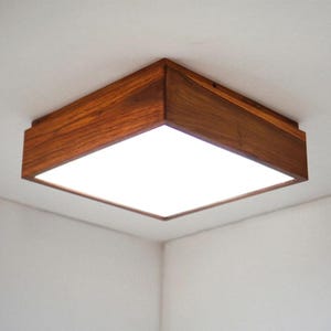 May include: A square ceiling light fixture featuring a dark brown wooden frame with a visible natural grain pattern. The center of the fixture consists of a flat, white, translucent panel that emits a bright, even light across the surface.