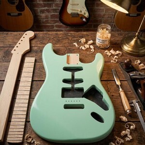 Könnte beinhalten: Ein hell mintgrüner E-Gitarrenkorpus mit gefrästen Aussparungen, ein separater, unbehandelter Ahornhals und ein Griffbrett. Holzspäne liegen verstreut daneben, zusammen mit einem Glas mit der Aufschrift Surf Green Finish und Werkzeugen.