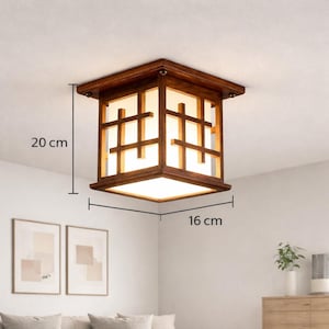 Könnte beinhalten: Eine quadratische Deckenleuchte aus Holz mit warmbraunem Finish. Die Leuchte hat ein gitterartiges Design mit einem weißen, beleuchteten Innenraum. Sie misst ca. 20 cm in der Höhe und 16 cm in der Breite. Das Licht ist an einer weißen Decke befestigt.
