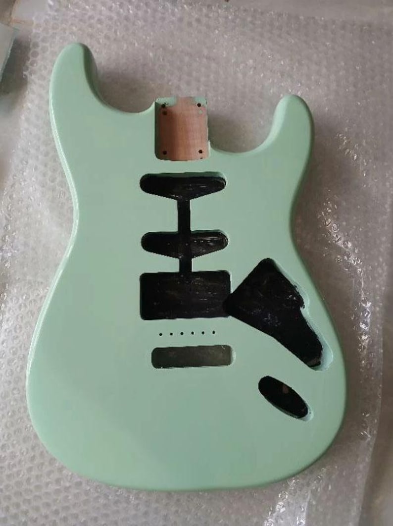 May include: A solid body electric guitar body finished in a light mint green color. The body features a double cutaway design with a natural wood neck pocket and several black-painted routed cavities for pickups, a bridge, and electronic controls.