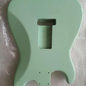 May include: A solid body electric guitar body finished in a smooth, light mint green color. The body features a central rectangular cavity for a pickup and a smaller rectangular area with four small pre-drilled holes for a bridge assembly.