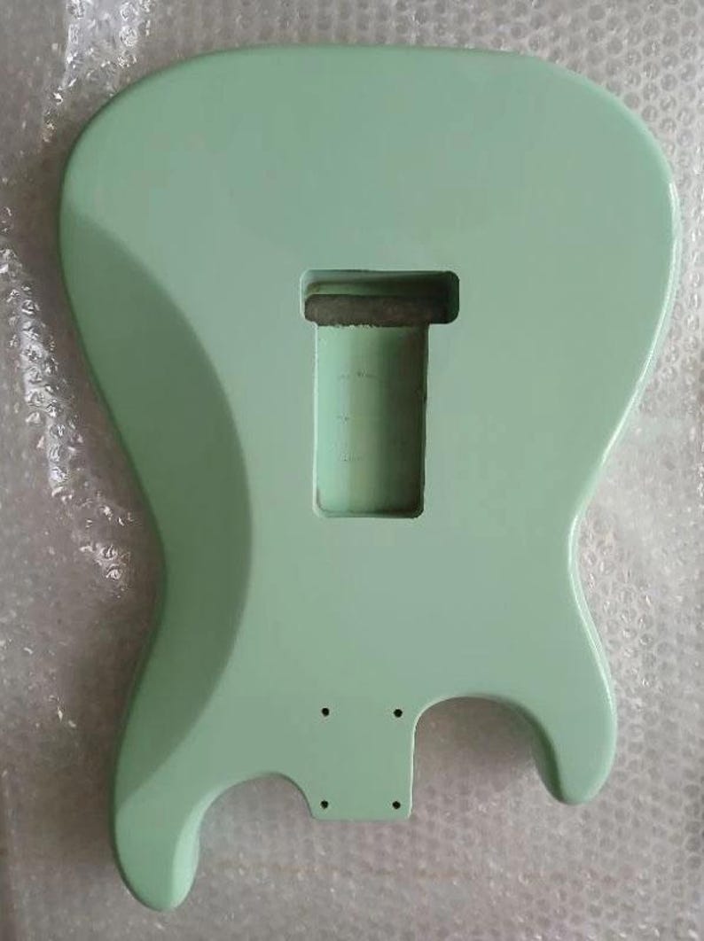 May include: A solid body electric guitar body finished in a smooth, light mint green color. The body features a central rectangular cavity for a pickup and a smaller rectangular area with four small pre-drilled holes for a bridge assembly.