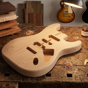 Könnte beinhalten: Ein unfertiger, heller E-Gitarrenkorpus aus Holz mit gefrästen Aussparungen für Tonabnehmer und Elektronik. Der Korpus hat eine glatte, geschliffene Oberfläche und liegt auf einer Werkbank neben Holzspänen, einem kleinen Glas mit bernsteinfarbener Beize und einem Messschieber.
