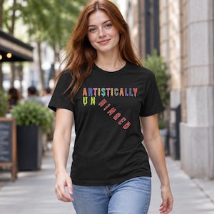 May include: A black short-sleeve t-shirt featuring the text ARTISTICALLY UNHINGED printed in a multi-coloured, dripping paint style. The word ARTISTICALLY is horizontal, while UNHINGED is positioned diagonally below it in red.