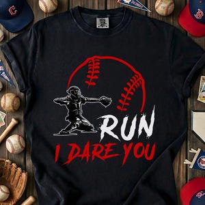 May include: Black t-shirt with a baseball-themed graphic. The design features a baseball with red stitching, a baseball player in a throwing pose, and the text "RUN I DARE YOU" in white and red. Baseball apparel.