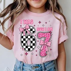 以下が含まれることがあります： 「I WAS 6 NOW I AM 7」の文字が入ったピンク色の半袖Tシャツ。数字の6は白黒の市松模様、数字の7はピンクのヒョウ柄。ピンクのリボンと星がシャツを飾っています。