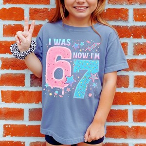 May include: A blue t-shirt with the text "I WAS 6 NOW I'M 7" in a colorful design. The number 6 is pink and donut-shaped, while the number 7 is blue with sprinkles. The shirt has a crew neck and short sleeves.