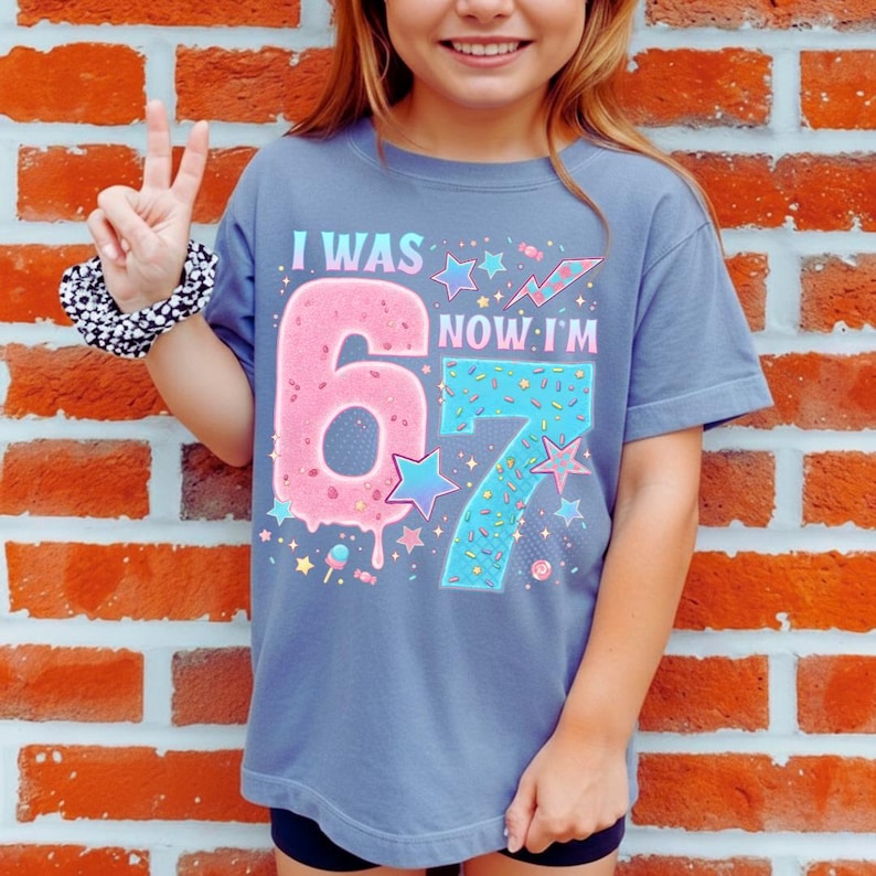 May include: A blue t-shirt with the text "I WAS 6 NOW I'M 7" in a colorful design. The number 6 is pink and donut-shaped, while the number 7 is blue with sprinkles. The shirt has a crew neck and short sleeves.