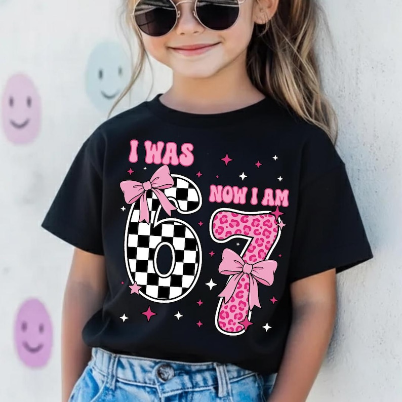 May include: Black t-shirt featuring the text "I WAS NOW I AM" in pink. The number "67" is displayed with a checkered pattern and leopard print, accented with pink bows and stars. A child is wearing the shirt.