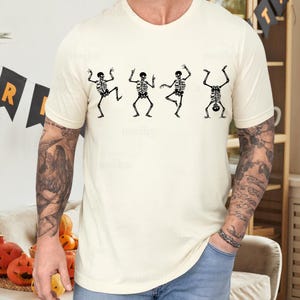 Pu&ograve; includere: T-shirt color crema con quattro figure di scheletri neri in varie pose di danza. Gli scheletri sono dettagliati con gabbie toraciche, colonne vertebrali e arti. La maglietta &egrave; uno stile casual, a maniche corte, adatto per Halloween o eventi a tema.