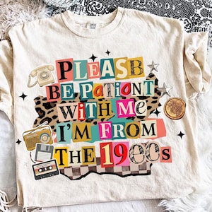 May include: Cream-colored t-shirt with the text "PLEASE BE PATIENT WITH ME I'M FROM THE 1900s" in a colorful, collage-style design. The design includes retro illustrations of a telephone, radio, floppy disk, cassette tape, and stars.