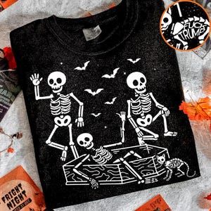 May include: Black Halloween sweatshirt with a white skeleton graphic. The design features three skeletons, bats, and a cat. One skeleton is in a coffin. The sweatshirt has the text "FUCK TRUMP" in a circle.
