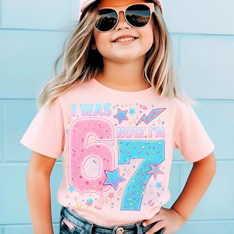 May include: A light pink t-shirt with the text "I WAS 6, NOW I'M 7". The number 6 is designed to look like a pink donut with sprinkles, and the number 7 is blue with sprinkles. The shirt is worn by a child.