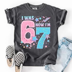 May include: A charcoal gray t-shirt with the text "I WAS 6, NOW I'M 7" in a colorful, playful design. The number 6 is pink and the number 7 is blue, with star and candy accents. Black and white checkered slip-on shoes and denim shorts are also visible.