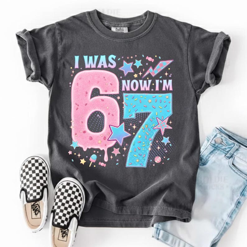 May include: A charcoal gray t-shirt with the text "I WAS 6, NOW I'M 7" in a colorful, playful design. The number 6 is pink and the number 7 is blue, with star and candy accents. Black and white checkered slip-on shoes and denim shorts are also visible.