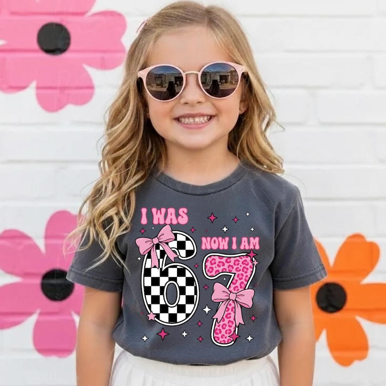 May include: A young girl wearing a grey t-shirt with the text "I WAS 67 NOW I AM" in pink. The number 6 is checkered, and the number 7 has a pink leopard print. Pink bows and stars decorate the shirt. The girl is wearing sunglasses.
