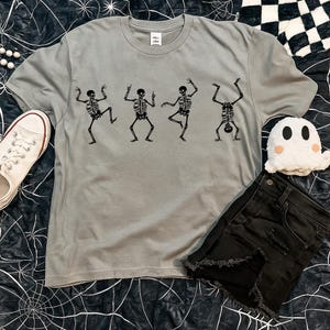 Pu&ograve; includere: Una maglietta grigia con una stampa nera di scheletri danzanti. La maglietta ha un girocollo e maniche corte. L'immagine mostra anche pantaloncini di jeans neri, una scarpa di tela bianca e un fantasma di peluche.
