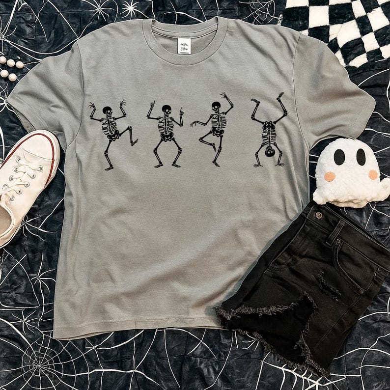 Pu&ograve; includere: Una maglietta grigia con una stampa nera di scheletri danzanti. La maglietta ha un girocollo e maniche corte. L'immagine mostra anche pantaloncini di jeans neri, una scarpa di tela bianca e un fantasma di peluche.