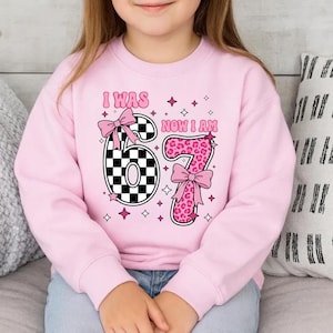 May include: A light pink sweatshirt featuring the text "I WAS 67 NOW I AM". The number 6 is a black and white checkerboard pattern, and the number 7 is pink leopard print. The design is decorated with pink bows and stars.