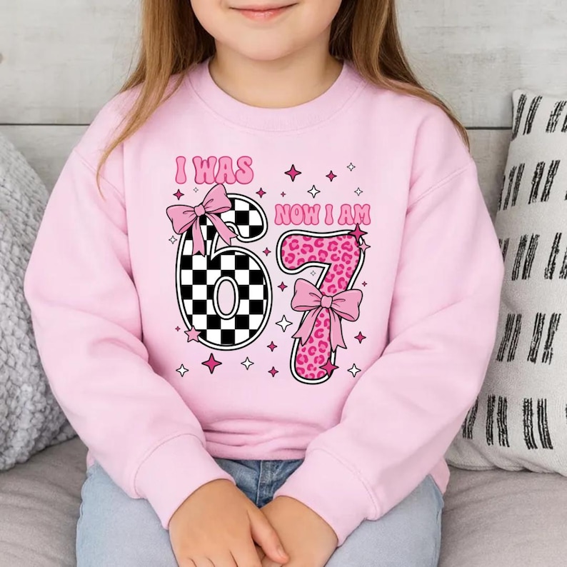 May include: A light pink sweatshirt featuring the text "I WAS 67 NOW I AM". The number 6 is a black and white checkerboard pattern, and the number 7 is pink leopard print. The design is decorated with pink bows and stars.