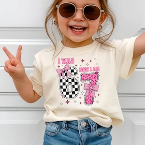 May include: A cream-coloured t-shirt with the text "I WAS NOW I AM 67". The number 6 is a black and white chequered pattern, and the number 7 is pink leopard print. Both numbers have pink bows.