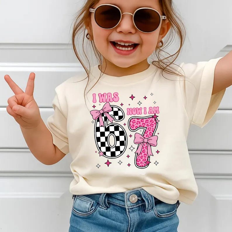May include: A cream-coloured t-shirt with the text "I WAS NOW I AM 67". The number 6 is a black and white chequered pattern, and the number 7 is pink leopard print. Both numbers have pink bows.