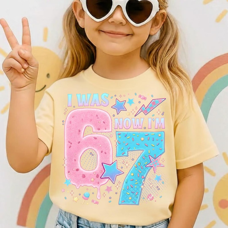 May include: A light yellow t-shirt with the text "I WAS 6 NOW I'M 7" in a colorful design. The number 6 is pink and the number 7 is blue. The shirt has a round neck and short sleeves.