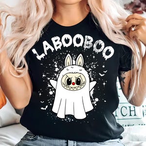Könnte beinhalten: Schwarzes T-Shirt mit dem weißen Text "LABOOBOO", das einen Cartoon-Geist-Charakter mit Hasenohren zeigt. Der Geist hat eine rote Nase und ein grinsendes Grinsen, mit weißen Spritzern und Fledermausgrafiken.