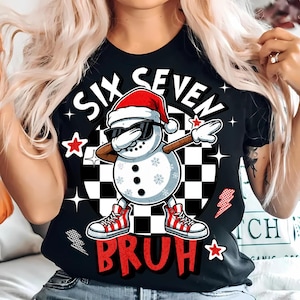 Może przedstawiać: Czarny t-shirt z grafiką bałwana w okularach przeciwsłonecznych i czapce Świętego Mikołaja, robiącego dabbing. Tekst na koszulce brzmi "SIX SEVEN BRUH" w kolorze białym i czerwonym. Bałwan stoi przed czarno-białym okręgiem w szachownicę.