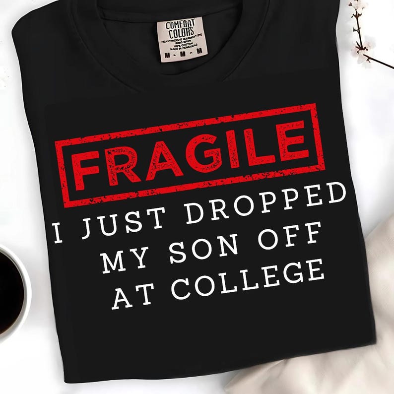 May include: Black t-shirt with the word "FRAGILE" in a red rectangle, and the text "I JUST DROPPED MY SON OFF AT COLLEGE" in white. The shirt has a crew neck and a Comfort Colors tag.