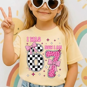 May include: A pale yellow t-shirt featuring the text "I WAS 67 NOW I AM" in pink. The number 67 is designed with a black and white checkered pattern and a pink bow. The number 7 has a pink leopard print and a pink bow.