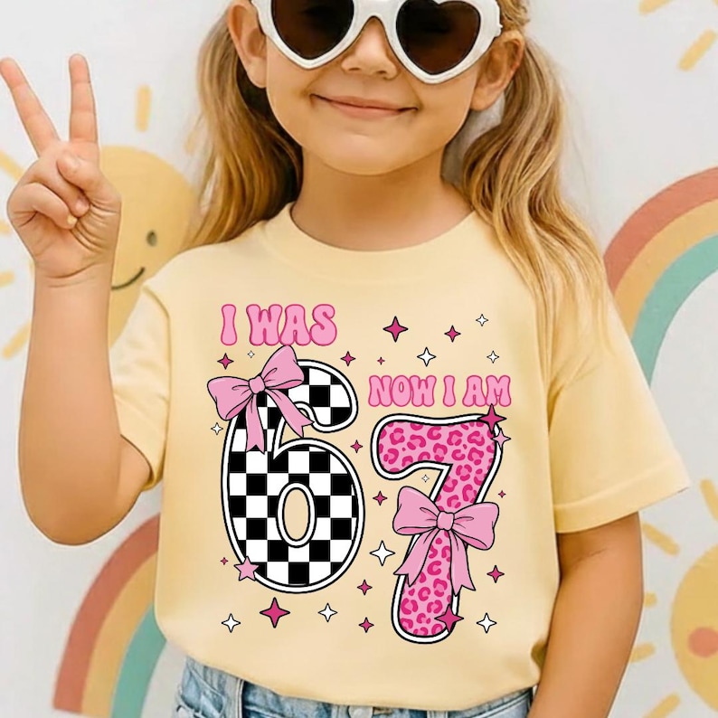May include: A pale yellow t-shirt featuring the text "I WAS 67 NOW I AM" in pink. The number 67 is designed with a black and white checkered pattern and a pink bow. The number 7 has a pink leopard print and a pink bow.