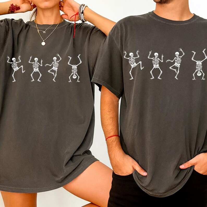 Pu&ograve; includere: Due magliette grigio scuro con un disegno di scheletro bianco. Gli scheletri sono raffigurati in posa di danza. Le magliette sono a maniche corte e hanno una vestibilit&agrave; ampia. L'immagine mostra le magliette indossate.