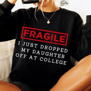 May include: Black sweatshirt with the word "FRAGILE" in a red rectangle, and the text "I JUST DROPPED MY DAUGHTER OFF AT COLLEGE" in white. The sweatshirt is a casual style.