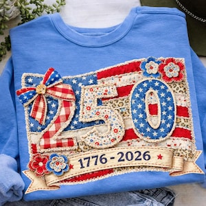 May include: Light blue sweatshirt with a patriotic design. Features a patchwork American flag with the number "250" and the years "1776-2026". Embellished with a bow, floral accents, and star details. A festive garment.