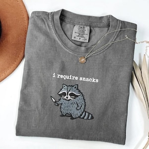 May include: Gray Comfort Colors t-shirt with the phrase "I require snacks" and a cartoon raccoon holding a knife. The shirt has a crew neck and short sleeves. A silver necklace with a pendant is draped across the shirt.
