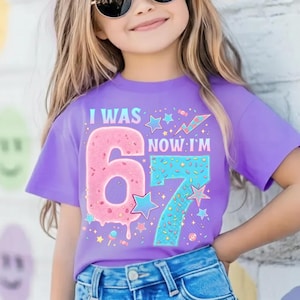 May include: A lilac-colored t-shirt with the text "I WAS 6 NOW I'M 7" in a colorful design. The number 6 is pink with sprinkles, and the number 7 is blue with sprinkles. Stars and candy graphics complete the design.