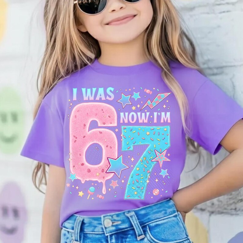 May include: A lilac-colored t-shirt with the text "I WAS 6 NOW I'M 7" in a colorful design. The number 6 is pink with sprinkles, and the number 7 is blue with sprinkles. Stars and candy graphics complete the design.