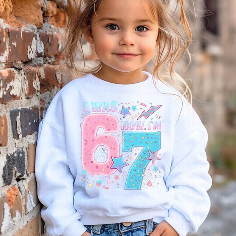 May include: A white sweatshirt with the text "I WAS 6, NOW I'M 7" in a colorful design. The number 6 is pink, and the number 7 is blue. The sweatshirt is for a child's birthday.
