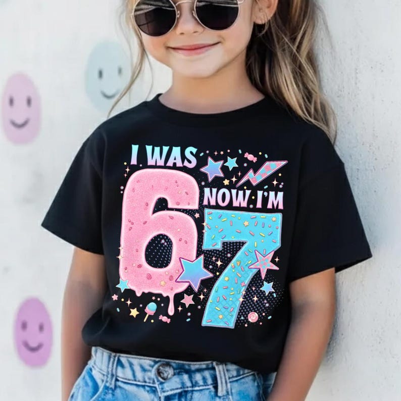 May include: Black t-shirt with the text "I WAS 6 NOW I'M 7". The number 6 is pink and the number 7 is blue, both decorated with stars and sprinkles. A fun birthday shirt for a child.