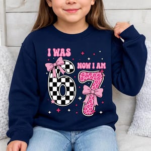May include: Navy blue sweatshirt with the text "I WAS 6 NOW I AM 7". The number 6 is designed with a black and white checkered pattern and a pink bow. The number 7 is pink with a leopard print and a pink bow.