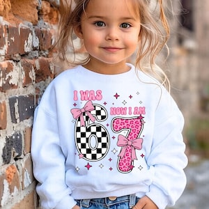 May include: White sweatshirt with the text "I WAS 6 NOW I AM 7". The number 6 has a black and white chequered pattern, and the number 7 has a pink leopard print. Pink bows and stars decorate the design.