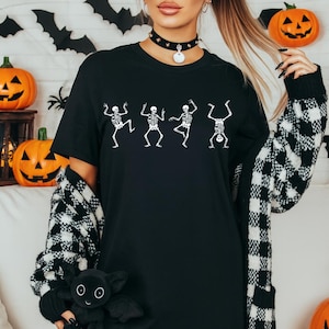 Pu&ograve; includere: T-shirt nero con grafica bianca di scheletri danzanti. L'immagine ha un tema di Halloween, con zucche e pipistrelli sullo sfondo. La modella indossa un choker nero e un cardigan a fantasia bianco e nero.