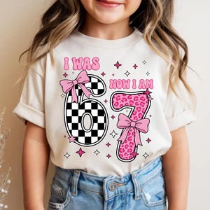 May include: A cream-coloured t-shirt with the text "I WAS 6, NOW I AM 7" in pink. The number 6 is black and white chequered, and the number 7 is pink leopard print. Pink bows and stars decorate the shirt.