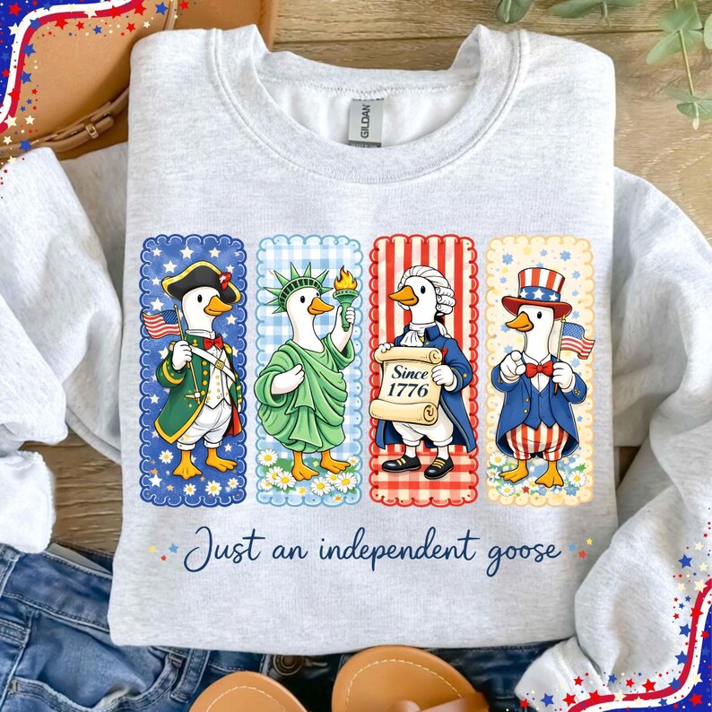May include: A light gray sweatshirt featuring four cartoon geese dressed in patriotic attire. Each goose represents a different historical figure, with the text "Just an independent goose" below the images. The design includes red, white, and blue colors.