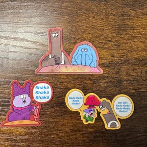 May include: A collection of colorful cartoon stickers featuring characters with speech bubbles. The speech bubbles contain phrases like "Shaka Shaka Shaka" and "Rada Rada Rada!!" on a wooden surface.